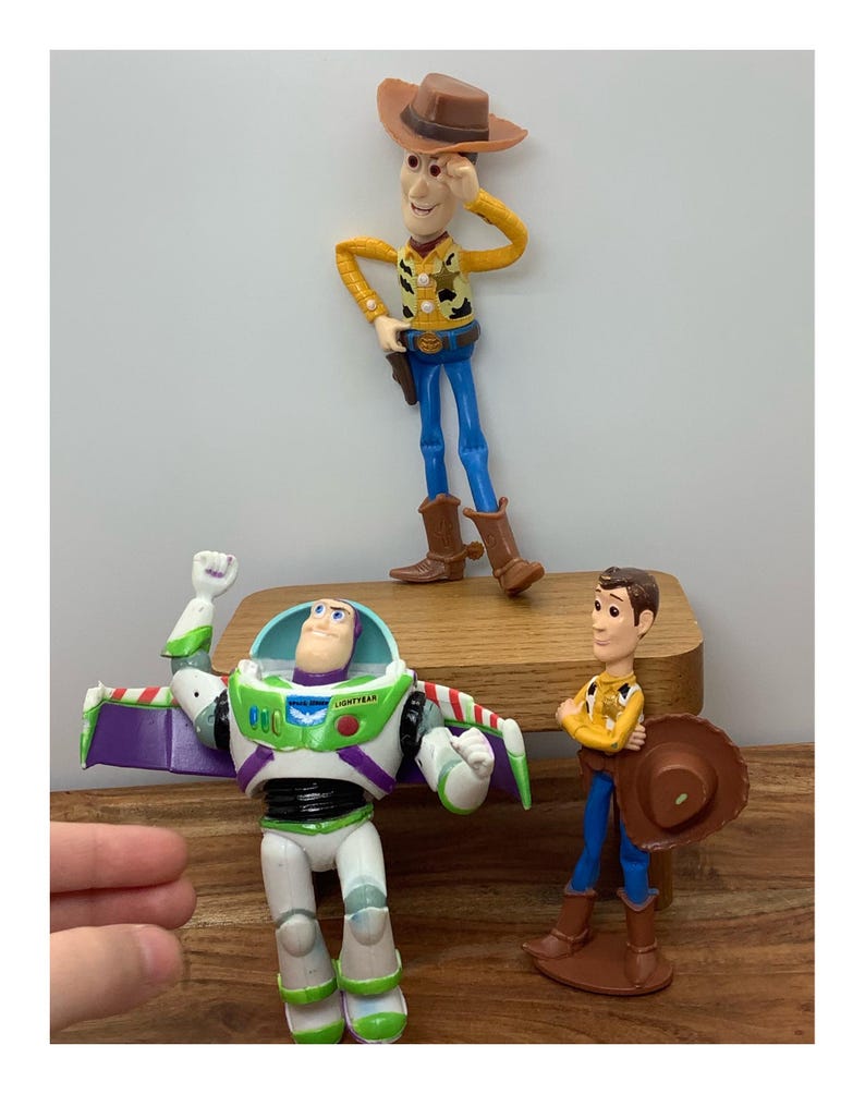 May include: Three Toy Story character figurines: Woody, Buzz Lightyear, and another Woody. Woody is wearing a cowboy hat, yellow shirt, blue trousers, and brown boots. Buzz Lightyear is in a white and green spacesuit. The other Woody is holding a hat.