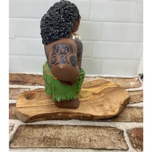 May include: A brown figurine of a person with dark curly hair, tattoos, and a green grass skirt. The figurine is posed on a wooden cutting board with a natural edge. The background is a white tiled wall.