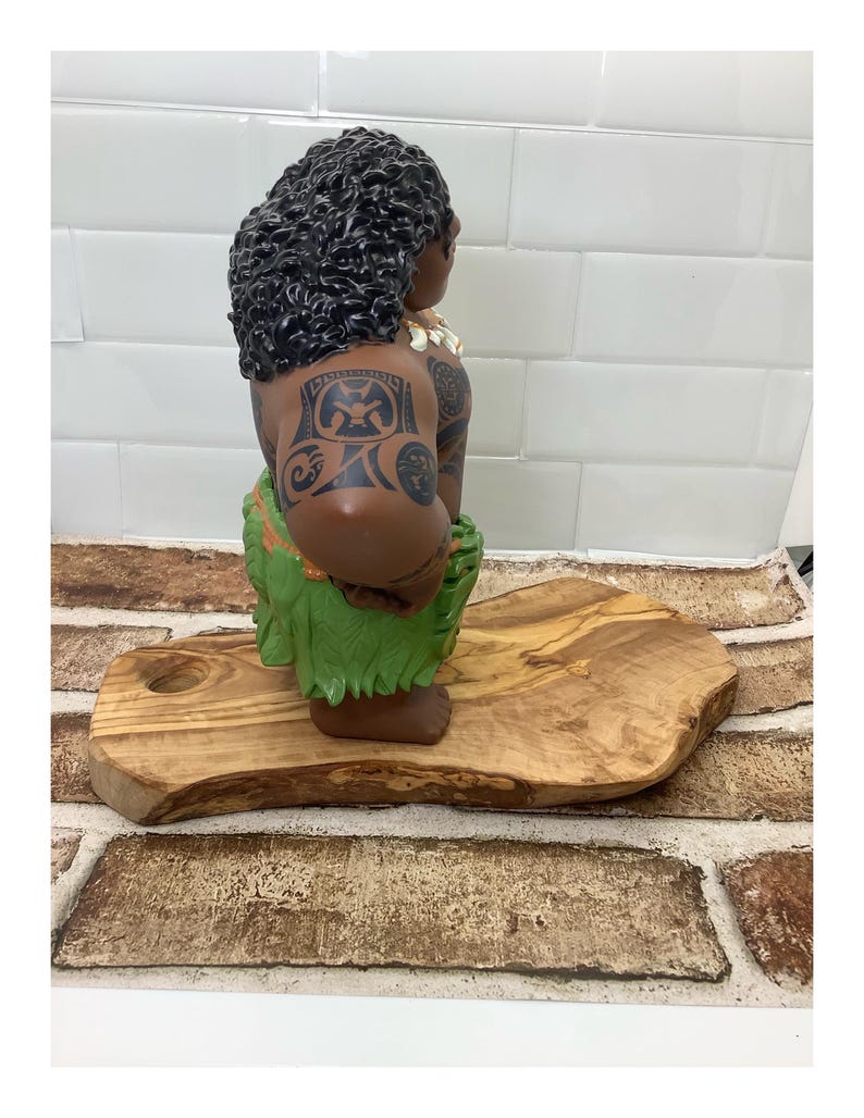 May include: A brown figurine of a person with dark curly hair, tattoos, and a green grass skirt. The figurine is posed on a wooden cutting board with a natural edge. The background is a white tiled wall.