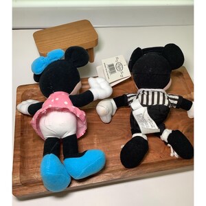 May include: Two plush Mickey Mouse and Minnie Mouse dolls. Minnie Mouse is wearing a pink polka dot dress, blue bow, and blue shoes. Mickey Mouse is wearing a black and white striped shirt. Both dolls have white gloves.