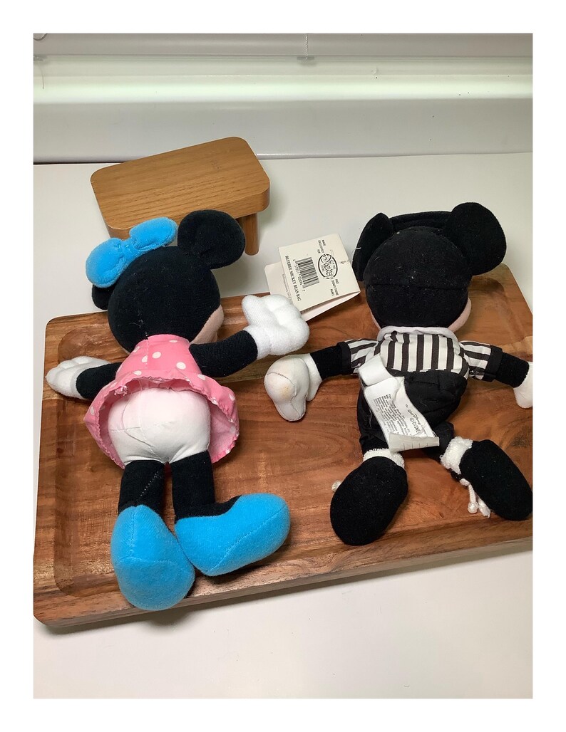 May include: Two plush Mickey Mouse and Minnie Mouse dolls. Minnie Mouse is wearing a pink polka dot dress, blue bow, and blue shoes. Mickey Mouse is wearing a black and white striped shirt. Both dolls have white gloves.