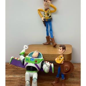 May include: Three Toy Story character figurines: Woody, Buzz Lightyear, and another Woody. Woody is wearing a cowboy hat, yellow shirt, blue trousers, and brown boots. Buzz Lightyear is in a white and green space suit. The other Woody is holding a hat.