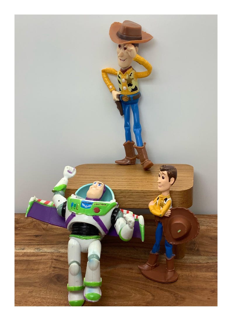 May include: Three Toy Story character figurines: Woody, Buzz Lightyear, and another Woody. Woody is wearing a cowboy hat, yellow shirt, blue trousers, and brown boots. Buzz Lightyear is in a white and green space suit. The other Woody is holding a hat.
