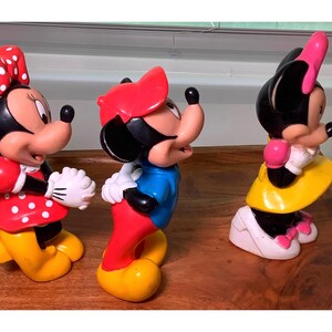 Pu&ograve; includere: Tre colorate statuette di Minnie Mouse. Le statuette sono in varie pose e abiti, tra cui abiti rossi a pois, gonne gialle e fiocchi rossi e rosa. Le statuette sono su una superficie di legno.