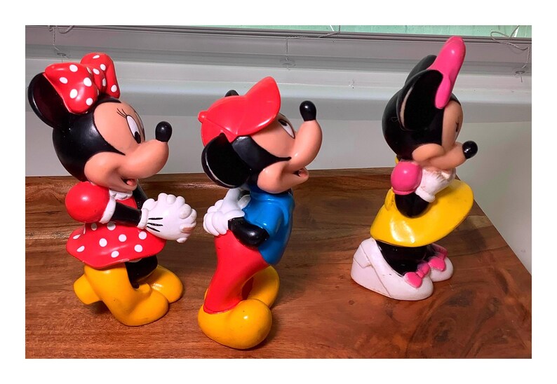 Pu&ograve; includere: Tre colorate statuette di Minnie Mouse. Le statuette sono in varie pose e abiti, tra cui abiti rossi a pois, gonne gialle e fiocchi rossi e rosa. Le statuette sono su una superficie di legno.
