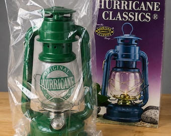 May include: A green metal hurricane lantern inside a clear plastic bag, positioned next to its original cardboard box. The box features the text HURRICANE CLASSICS and displays an image of a blue lantern. The lantern has a clear glass globe.