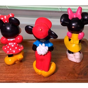 Pu&ograve; includere: Tre statuette vintage di Topolino e Minnie. La statuetta di Topolino indossa un berretto rosso, una camicia blu e pantaloni rossi. Le statuette di Minnie indossano abiti rossi a pois bianchi e abiti gialli con fiocchi rosa.