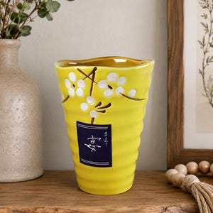 May include: A bright yellow ceramic vase with a ribbed, tapered body. The vase features a hand-painted design of white flowers on brown stems and a rectangular dark blue label with white Japanese characters centered on the front surface.