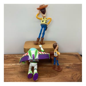 May include: Three Toy Story character figurines: Woody in a cowboy hat, Buzz Lightyear with wings, and a smaller Woody figure. The toys are on a wooden surface, with a neutral background. The colours are vibrant, with blue, yellow, green, and purple.