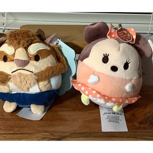 May include: Two plush toys, one resembling the Beast from Beauty and the Beast, the other a character with a bow. The Beast has brown fur and blue clothing. The other character is peach-colored with a polka-dot skirt.