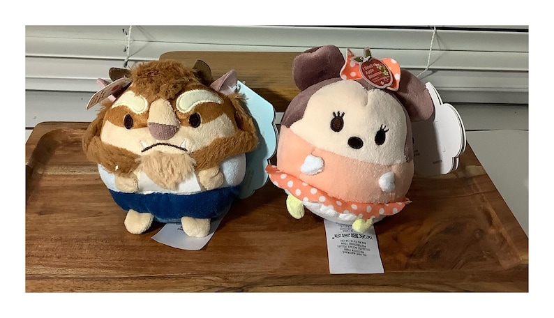May include: Two plush toys, one resembling the Beast from Beauty and the Beast, the other a character with a bow. The Beast has brown fur and blue clothing. The other character is peach-colored with a polka-dot skirt.