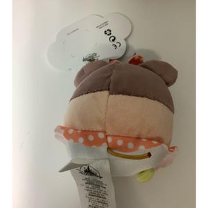 May include: A plush toy featuring a character with a brown head, peach face, and orange polka-dot trim. The toy has a white tag with text and a Disney Store logo. The toy is on a white background.