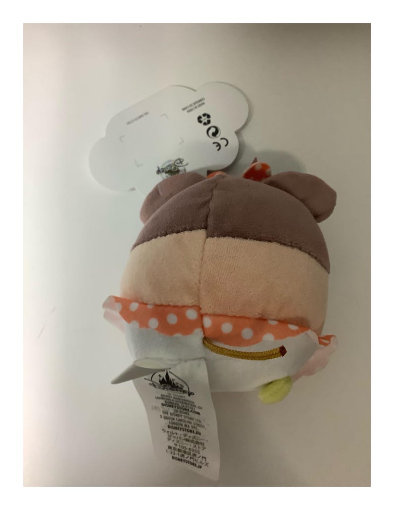 May include: A plush toy featuring a character with a brown head, peach face, and orange polka-dot trim. The toy has a white tag with text and a Disney Store logo. The toy is on a white background.