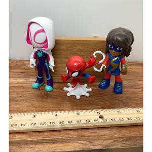 Op de afbeelding: Drie superheldenbeeldjes: Spider-Gwen in wit en paars, een rode Spider-Man en Ms. Marvel in blauw en goud. De figuren staan op een houten oppervlak, met een liniaal die centimeters aangeeft.