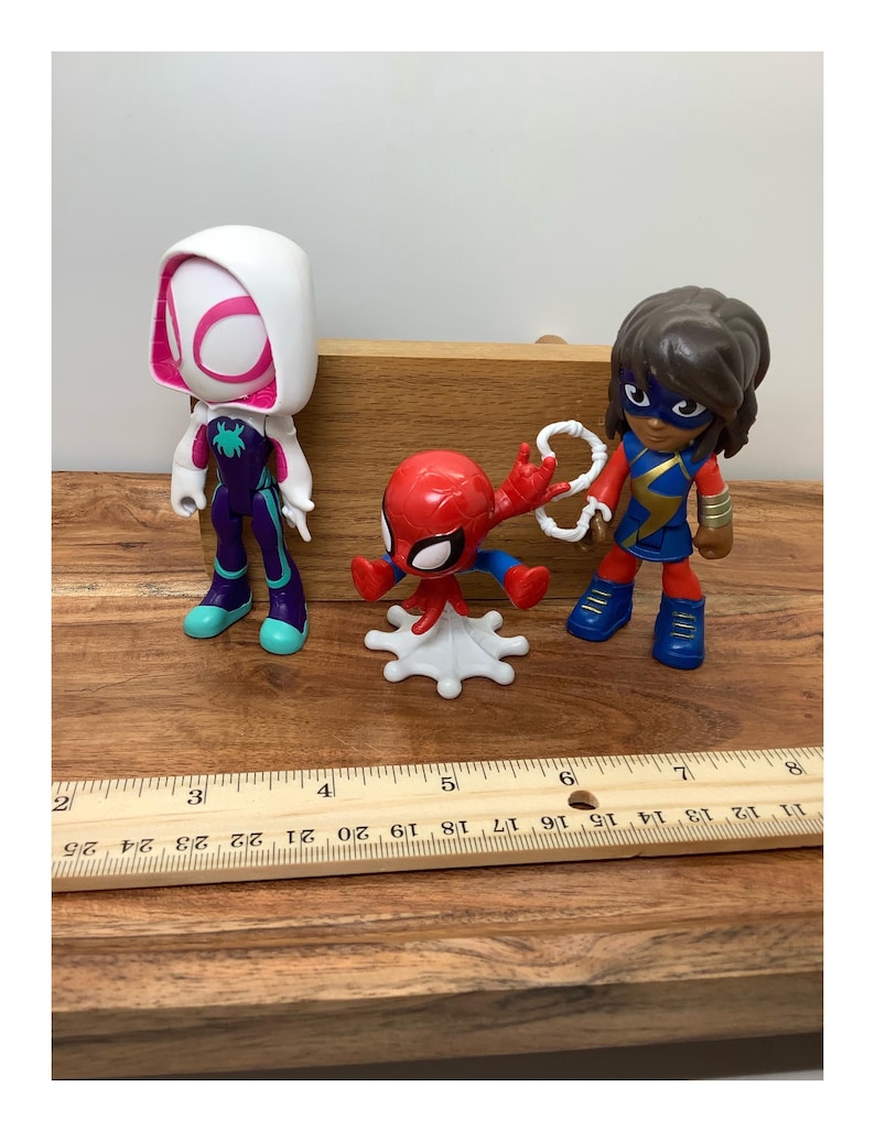 Op de afbeelding: Drie superheldenbeeldjes: Spider-Gwen in wit en paars, een rode Spider-Man en Ms. Marvel in blauw en goud. De figuren staan op een houten oppervlak, met een liniaal die centimeters aangeeft.