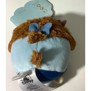May include: A plush toy featuring a brown, furry head with blue bow accents and a light blue body. The toy has a Disney tag attached. The plush is designed in a rounded shape.