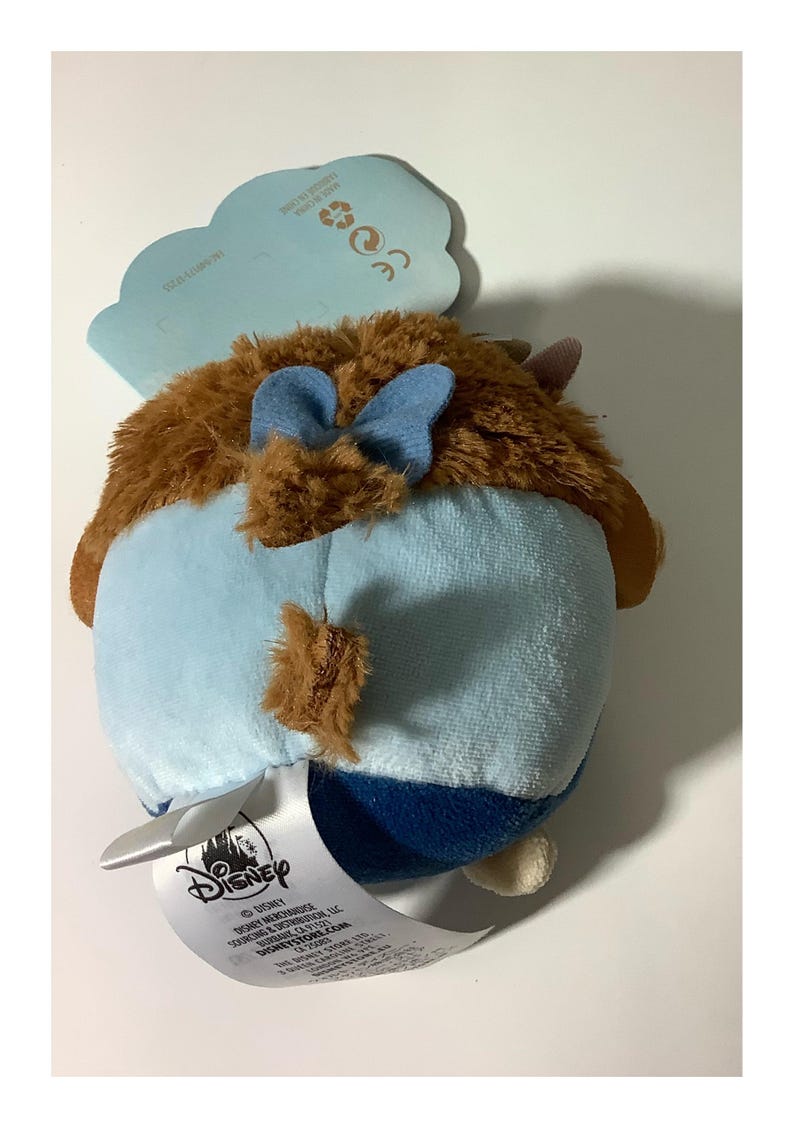 May include: A plush toy featuring a brown, furry head with blue bow accents and a light blue body. The toy has a Disney tag attached. The plush is designed in a rounded shape.