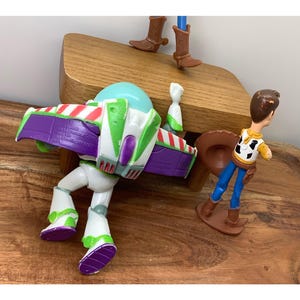 May include: Toy Story character figures of Buzz Lightyear and Woody. Buzz is white, green, purple, and red. Woody wears a yellow shirt, blue trousers, and a brown cowboy hat. The figures are on a wooden surface.
