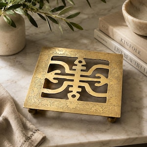May include: Square brass trivet with an ornate, openwork design. The trivet features a central geometric pattern and floral border. It rests on four small, rounded feet. The background includes books, a marble bowl, and a vase with greenery.