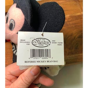 May include: A Referee Mickey Bean Bag plush toy. The plush toy has black ears, a black head, and a pink face. The tag reads "MADE EXCLUSIVELY FOR THE DISNEY STORE AND DISNEY THEME PARKS" and "REFEREE MICKEY BEAN BAG."