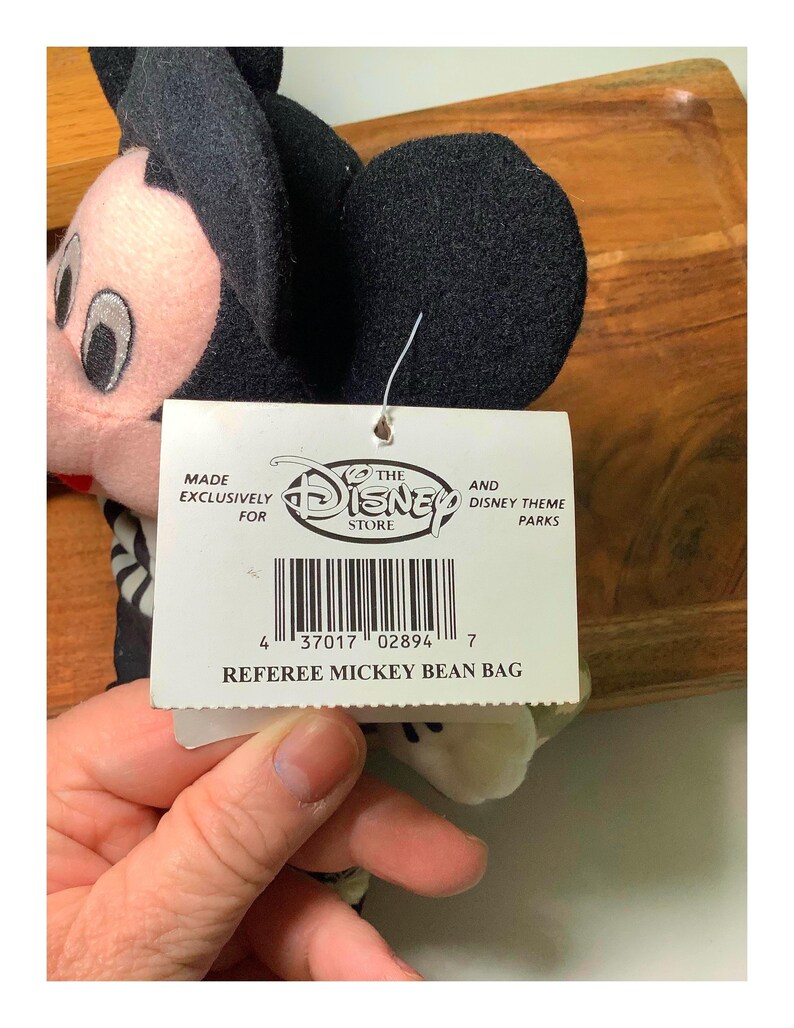 May include: A Referee Mickey Bean Bag plush toy. The plush toy has black ears, a black head, and a pink face. The tag reads "MADE EXCLUSIVELY FOR THE DISNEY STORE AND DISNEY THEME PARKS" and "REFEREE MICKEY BEAN BAG."