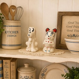 May include: A shelf displays kitchen decor, including a ceramic jar with "KITCHEN THINGS" text, wooden spoons, and a whisk. Two vintage ceramic figurines, a dog and a bear, sit on a doily. A framed print reads "Good Food, Good Friends, Good Times."