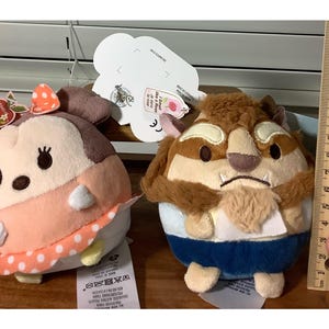 May include: Two plush character toys. One is a light pink and white character with a polka dot bow. The other is a brown and tan character with a blue outfit. A ruler shows the toys are approximately 4 inches tall.