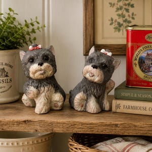 May include: Two small ceramic figurines of Yorkshire Terrier dogs with dark gray and tan fur, each wearing a tiny red and white bow on its head. The dogs are sitting in a playful pose with their heads tilted, featuring large dark eyes and noses.