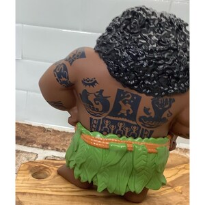 May include: A figurine of a muscular character with dark skin, curly black hair, and detailed tattoos on the back. The character wears a green leaf skirt with an orange waistband. The figurine is posed on a wooden cutting board.