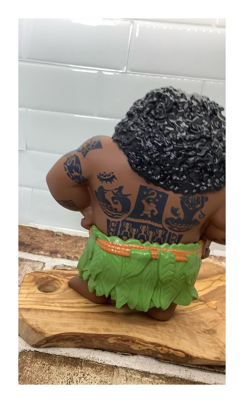 May include: A figurine of a muscular character with dark skin, curly black hair, and detailed tattoos on the back. The character wears a green leaf skirt with an orange waistband. The figurine is posed on a wooden cutting board.