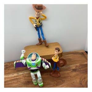 May include: Three Toy Story character figurines: Woody, Buzz Lightyear, and a third character. Woody is in a cowboy hat and Buzz has wings. The figurines are on a wooden surface.