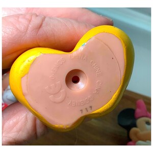 Pu&ograve; includere: Primo piano di un giocattolo giallo a forma di cuore con una base rosa chiaro. La base ha il testo "CHINA" e "DISNEY" stampato sopra. Il giocattolo &egrave; tenuto in una mano.