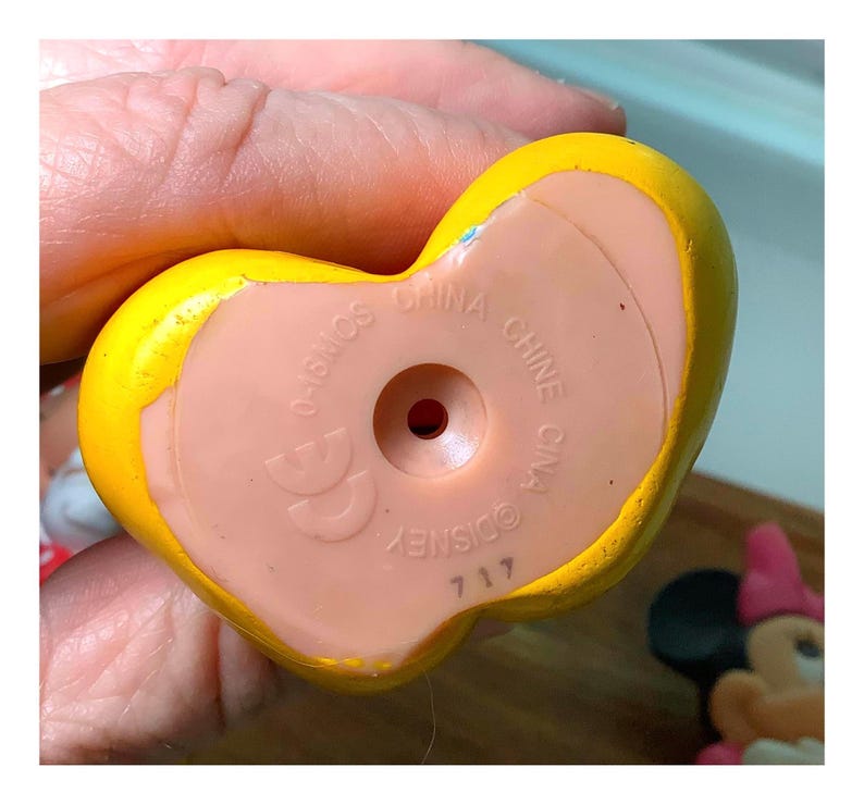 Pu&ograve; includere: Primo piano di un giocattolo giallo a forma di cuore con una base rosa chiaro. La base ha il testo "CHINA" e "DISNEY" stampato sopra. Il giocattolo &egrave; tenuto in una mano.