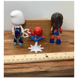 Op de afbeelding: Drie superheldenactiefiguren: Spider-Gwen, Spider-Man en Ms. Marvel. Spider-Man staat in een dynamische pose, terwijl de anderen rechtop staan. De figuren staan op een houten oppervlak met een liniaal.