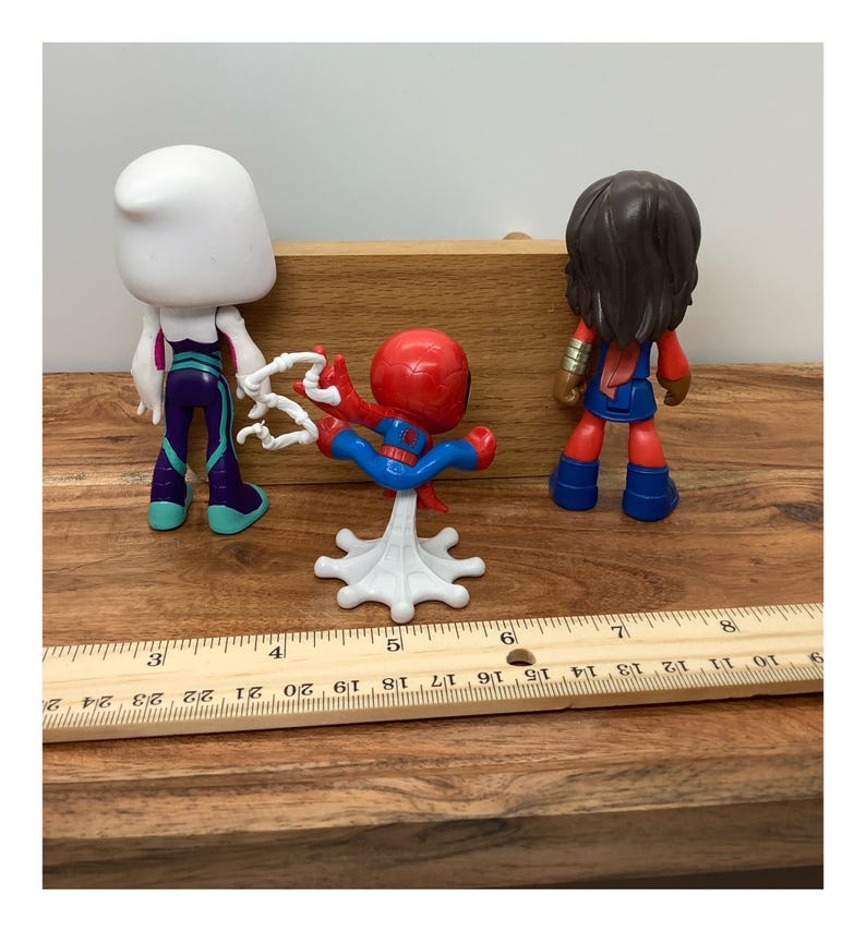 Op de afbeelding: Drie superheldenactiefiguren: Spider-Gwen, Spider-Man en Ms. Marvel. Spider-Man staat in een dynamische pose, terwijl de anderen rechtop staan. De figuren staan op een houten oppervlak met een liniaal.