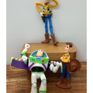 May include: Three Toy Story character figurines: Woody, Buzz Lightyear, and another Woody. Woody wears a cowboy hat, yellow shirt, blue trousers, and brown boots. Buzz Lightyear is in a white and green spacesuit. The other Woody is holding a hat.