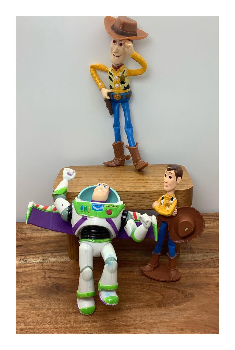 May include: Three Toy Story character figurines: Woody, Buzz Lightyear, and another Woody. Woody wears a cowboy hat, yellow shirt, blue trousers, and brown boots. Buzz Lightyear is in a white and green spacesuit. The other Woody is holding a hat.