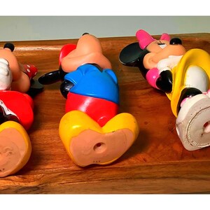 Pu&ograve; includere: Tre statuette vintage di Topolino e Minnie in un vassoio di legno. Le figure sono sdraiate, mostrando i loro abiti colorati: rosso, blu, giallo e rosa. Le statuette sono realizzate in un materiale duro e hanno un foro sul fondo.