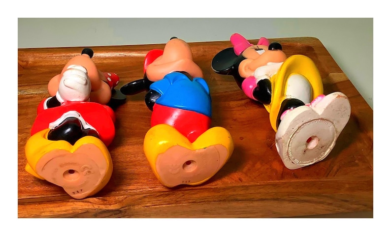 Pu&ograve; includere: Tre statuette vintage di Topolino e Minnie in un vassoio di legno. Le figure sono sdraiate, mostrando i loro abiti colorati: rosso, blu, giallo e rosa. Le statuette sono realizzate in un materiale duro e hanno un foro sul fondo.
