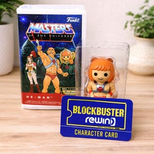 May include: A Funko Masters of the Universe collectible figure in a clear plastic case, with a Blockbuster Rewind character card. The figure is of a character with orange hair and a red and blue outfit. The box features He-Man.