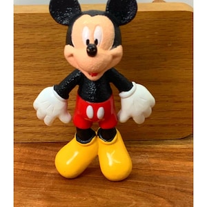May include: A figurine of Mickey Mouse, a classic cartoon character. Mickey has black ears, a black torso, red shorts, white gloves, and yellow shoes. The figurine is standing with arms outstretched.