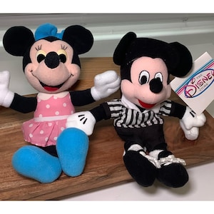 May include: Two plush Disney character dolls, Minnie Mouse and Mickey Mouse, are displayed. Minnie wears a pink polka-dot dress and blue bow. Mickey is in a black and white striped shirt. A Disney Store tag is visible.