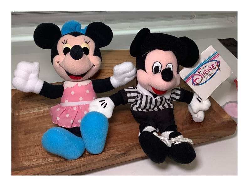 May include: Two plush Disney character dolls, Minnie Mouse and Mickey Mouse, are displayed. Minnie wears a pink polka-dot dress and blue bow. Mickey is in a black and white striped shirt. A Disney Store tag is visible.