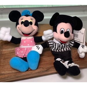 May include: Two plush Disney character bean bag dolls. Minnie Mouse wears a pink polka dot dress and blue shoes. Mickey Mouse is dressed in a black and white striped shirt and black pants. Both have white gloves and black ears.