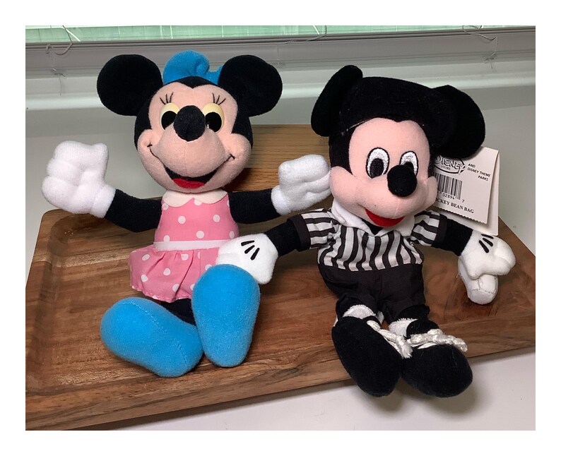May include: Two plush Disney character bean bag dolls. Minnie Mouse wears a pink polka dot dress and blue shoes. Mickey Mouse is dressed in a black and white striped shirt and black pants. Both have white gloves and black ears.