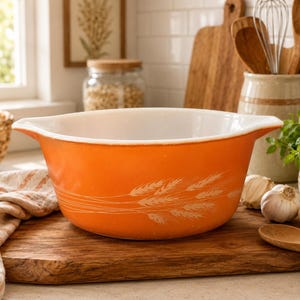 May include: An orange and white vintage mixing bowl with a wheat design. The bowl is sitting on a wooden cutting board with a wooden spoon and garlic cloves. Kitchenware and home decor.