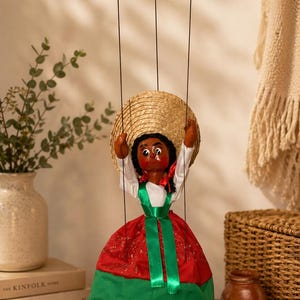 May include: A handcrafted marionette doll dressed in a traditional Mexican outfit. The doll has a straw hat, a red and green dress with a white lace trim, and is suspended by strings from a wooden bar. The doll's face is painted with dark features.