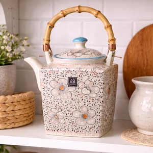 May include: Square ceramic teapot with a bamboo handle and floral pattern. The teapot features a light blue lid and a spout. The floral design includes orange and blue accents. The teapot is displayed on a white shelf.