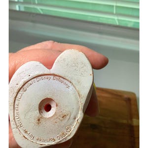 Pu&ograve; includere: Primo piano della base di un giocattolo vintage in plastica bianca. La base &egrave; a forma di cuore con un foro rosso al centro. Il testo recita "Walt Disney Company" e "&copy;1986 Tommee Tippee, Northvale, N.J."