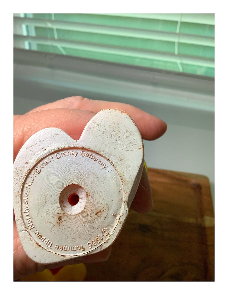 Pu&ograve; includere: Primo piano della base di un giocattolo vintage in plastica bianca. La base &egrave; a forma di cuore con un foro rosso al centro. Il testo recita "Walt Disney Company" e "&copy;1986 Tommee Tippee, Northvale, N.J."